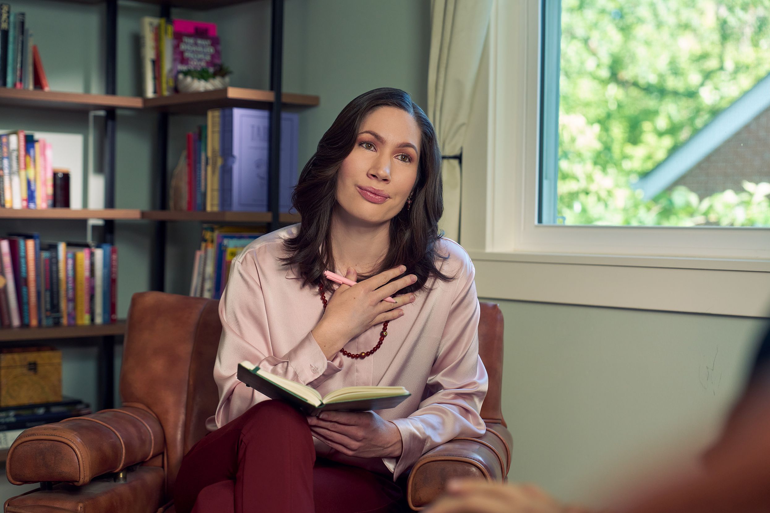 An Eastern European white woman therapist sits in a large chair and holds her hand up to her chest with an understanding look on her face. She is holding a pen and open notebook, and is looking at a client off camera. The therapist's office has several books on display and a window letting in natural light. The therapist is wearing a silk pink top, maroon dress pants, and a minimalist necklace.