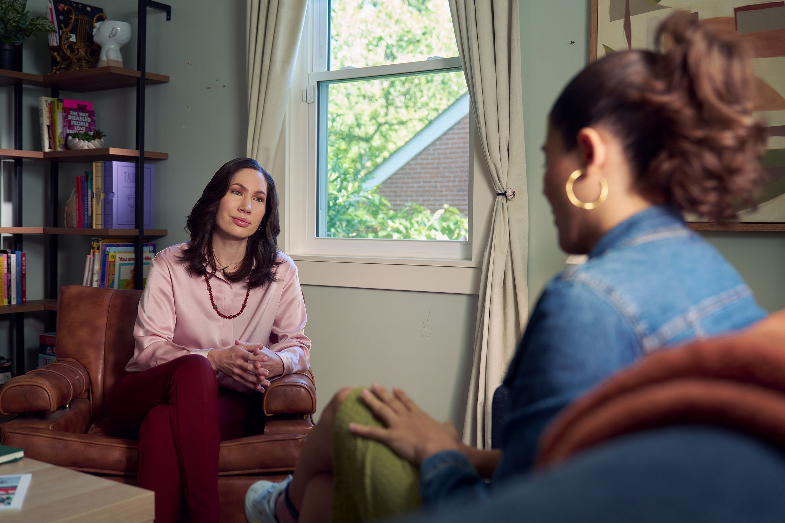 An Eastern European white woman therapist sits in a large chair and looks at a client seated on a couch in front of her. The client is a Latina woman with dark brown wavy hair pulled into a ponytail. The therapist's office has several books on display, an abstract art piece, and a window letting in natural light. The therapist is wearing a silk pink top, maroon dress pants, and a minimalist necklace. The client is wearing gold hoop earrings, a jean jacket, green skirt, and sneakers.