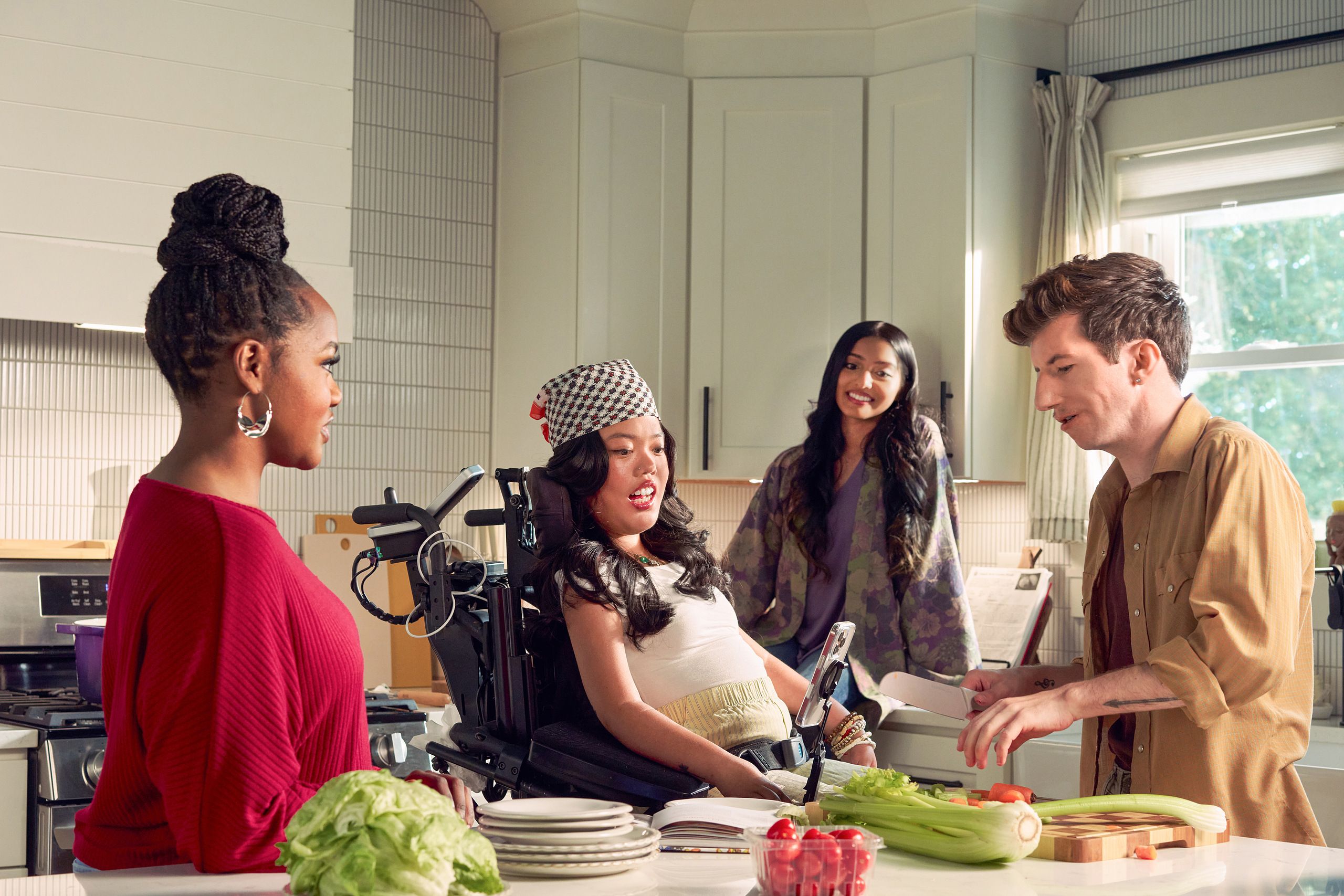Four roommates hang out in their bright kitchen. One roommate, a white male with Moebius Syndrome and a face that is paralyzed, chops vegetables. Another roomate, a SouthEast Asian woman with curled black hair seated in her power wheelchair, watches intently. A third roommate with dark hair wearing a purple floral dress sits on a counter and smiles while watching him prepare the vegetables. A fourth roommate, a black female with a braided bun wearing a maroon top, chats with him.