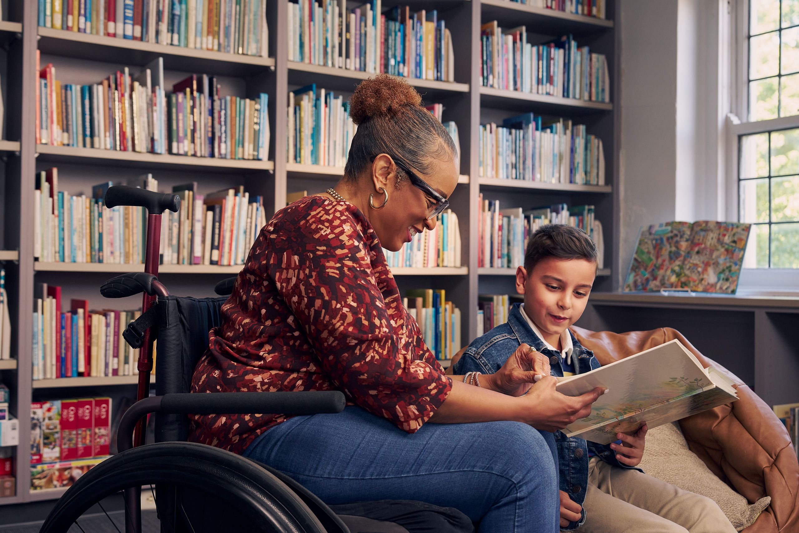 A young boy sitting in a bean bag chair and his African American teacher seated in a manual wheelchair hold and look at a picture book together. The teacher has a red walking cane fastened to the back of her wheelchair. She has medium brown skin, reddish brown hair pulled into a short natural ponytail, and is wearing reading glasses, a red patterned blouse, and jeans. The young student is a Hispanic and Mediterranean-American boy with short brown hair. He has olive skin and is wearing a blue striped shirt, a jean jacket, and khaki pants. A wall of children's books are behind them.