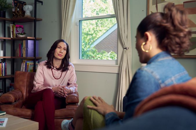 An Eastern European white woman therapist sits in a large chair and looks at a client seated on a couch in front of her. The client is a Latina woman with dark brown wavy hair pulled into a ponytail. The therapist's office has several books on display, an abstract art piece, and a window letting in natural light. The therapist is wearing a silk pink top, maroon dress pants, and a minimalist necklace. The client is wearing gold hoop earrings, a jean jacket, green skirt, and sneakers.