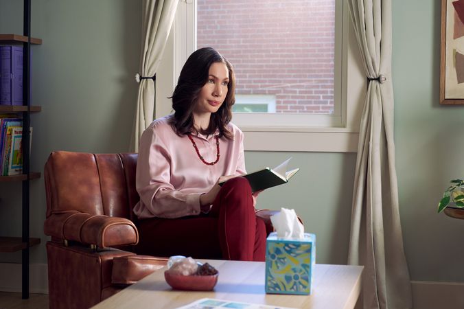 An Eastern European white woman therapist sits in a large chair looks at a client off camera. She is holding an open notebook. A tissue box and a red marble heasrt-shaped bowl of crystals are on a low coffee table in front of her. The therapist's office has several books on display and a window letting in natural light. The therapist is wearing a silk pink top, maroon dress pants, and a minimalist necklace.