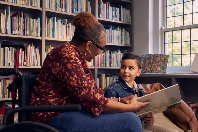 A young boy sitting in a bean bag chair looks up at his teacher, an African American woman seated in a manual wheelchair. They are both holding a picture book. The teacher has a red walking cane fastened to the back of her wheelchair. She has medium brown skin, reddish brown hair pulled into a short natural ponytail, and is wearing reading glasses, a red patterned blouse, and jeans. The young student is a Hispanic and Mediterranean-American boy with short brown hair. He  has olive skin and is wearing a blue striped shirt, a jean jacket, and khaki pants. A wall of children's books are behind them.